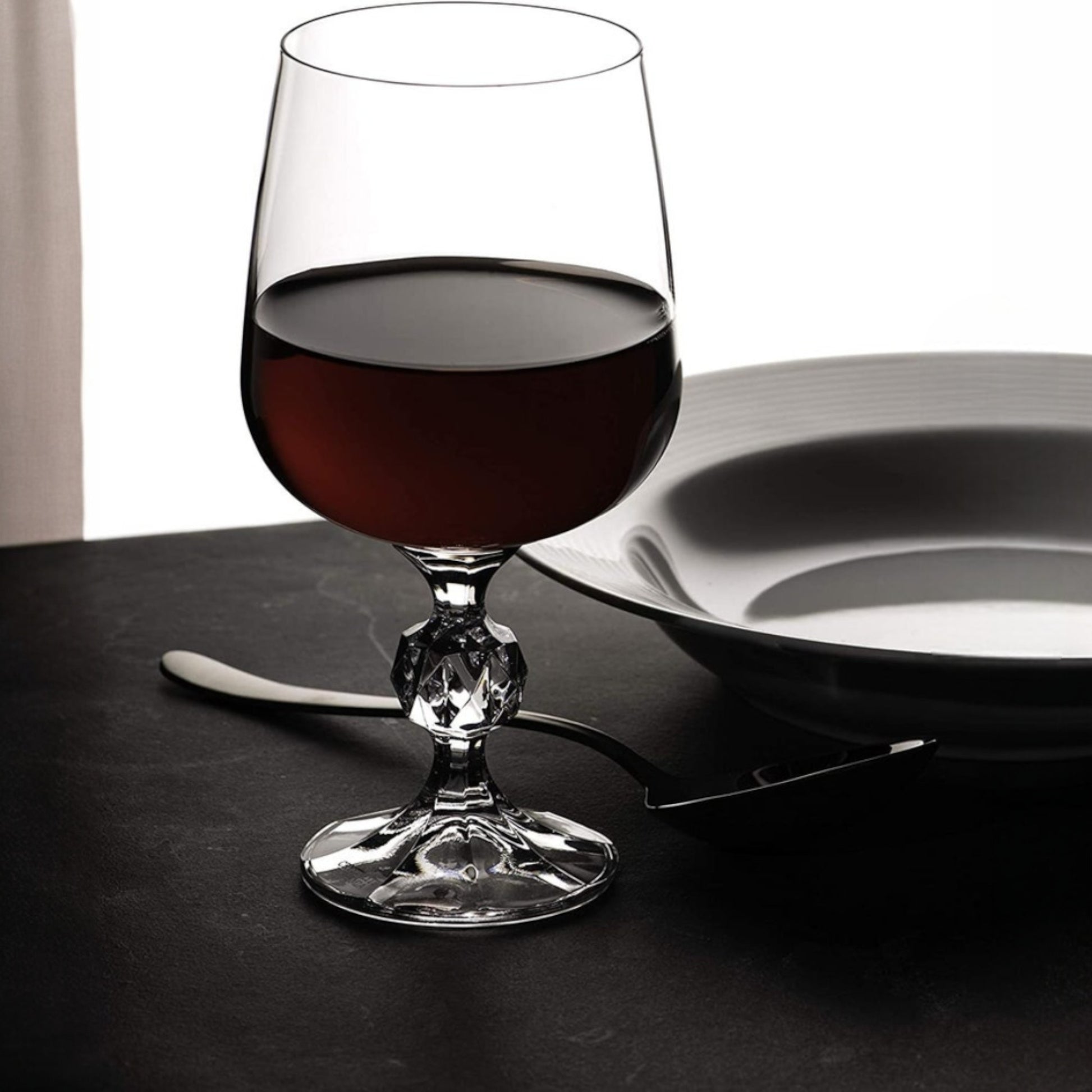 Glass of red wine on a dark surface with a white plate and cutlery.