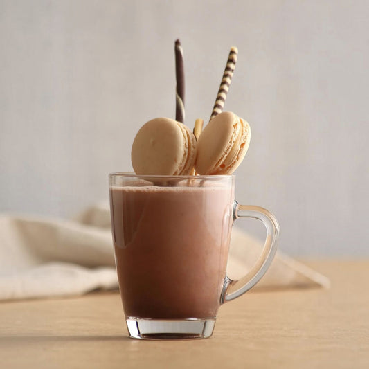 A clear glass mug filled with a brown chocolate drink, set on a light wooden surface against a soft neutral background. Two macarons and two striped straws are placed on the rim of the mug.