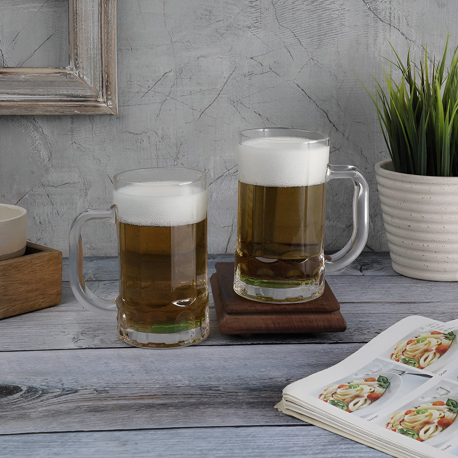 Two glasses of beer on a wooden table with a plant and framed picture in the background.
